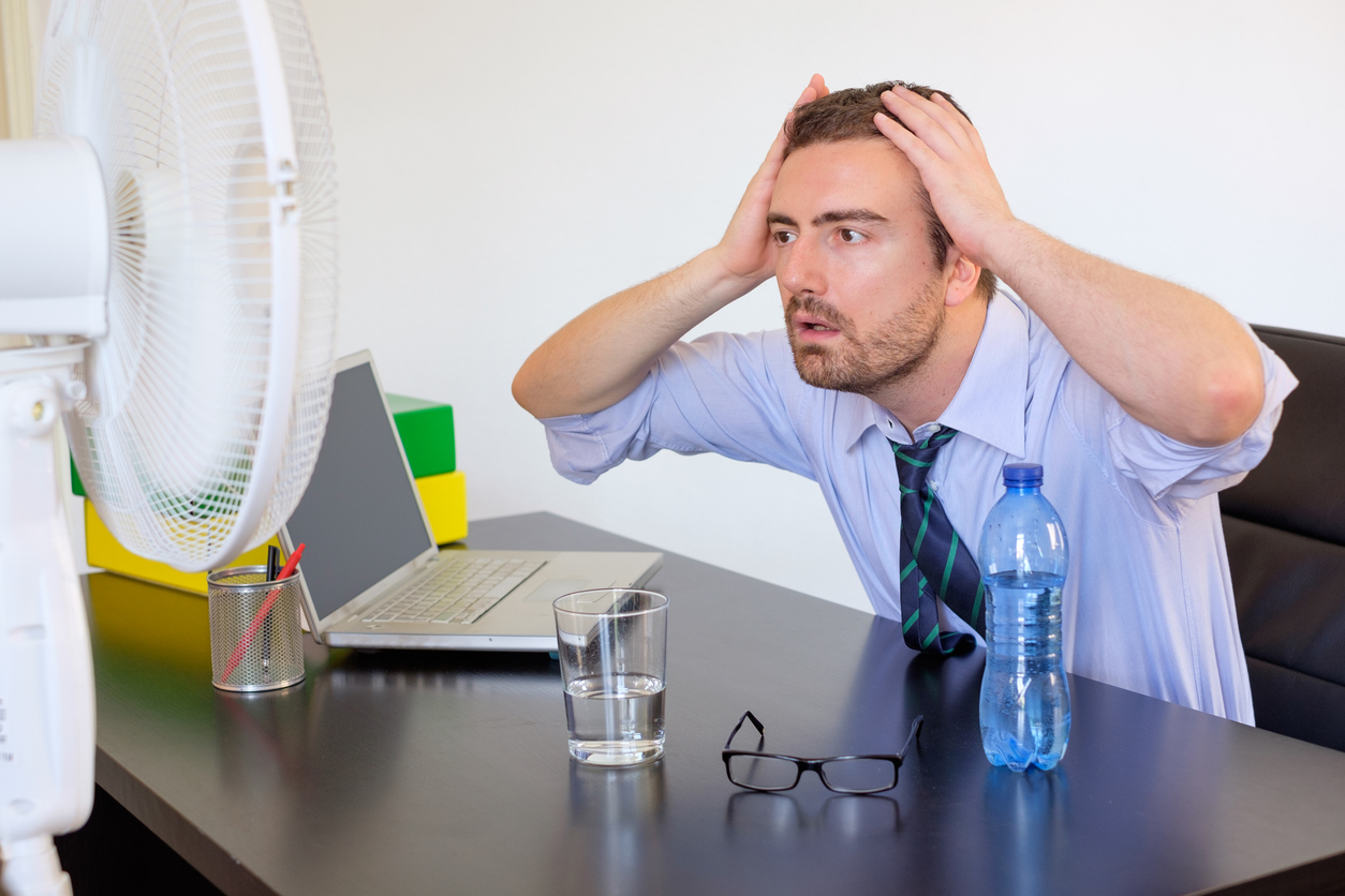 Man sitting at a desk in a suit and his hands on his head with a fan blowing on him.