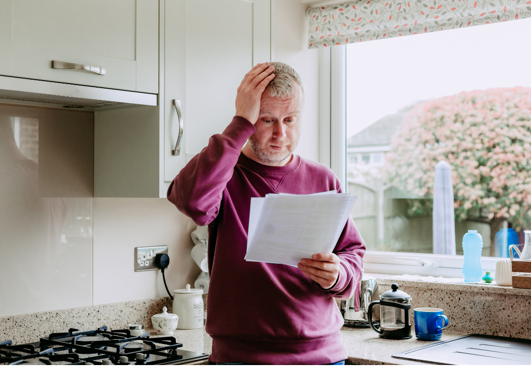 Man standing in kitchen, looking at energy bill with a stressed demeanor. Hand on head.
